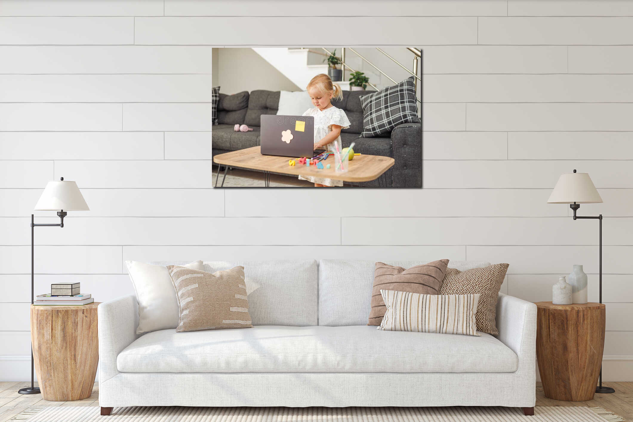 Young blond European girl using laptop at table with colored pencils in modern living room with gray sofa interior mockup